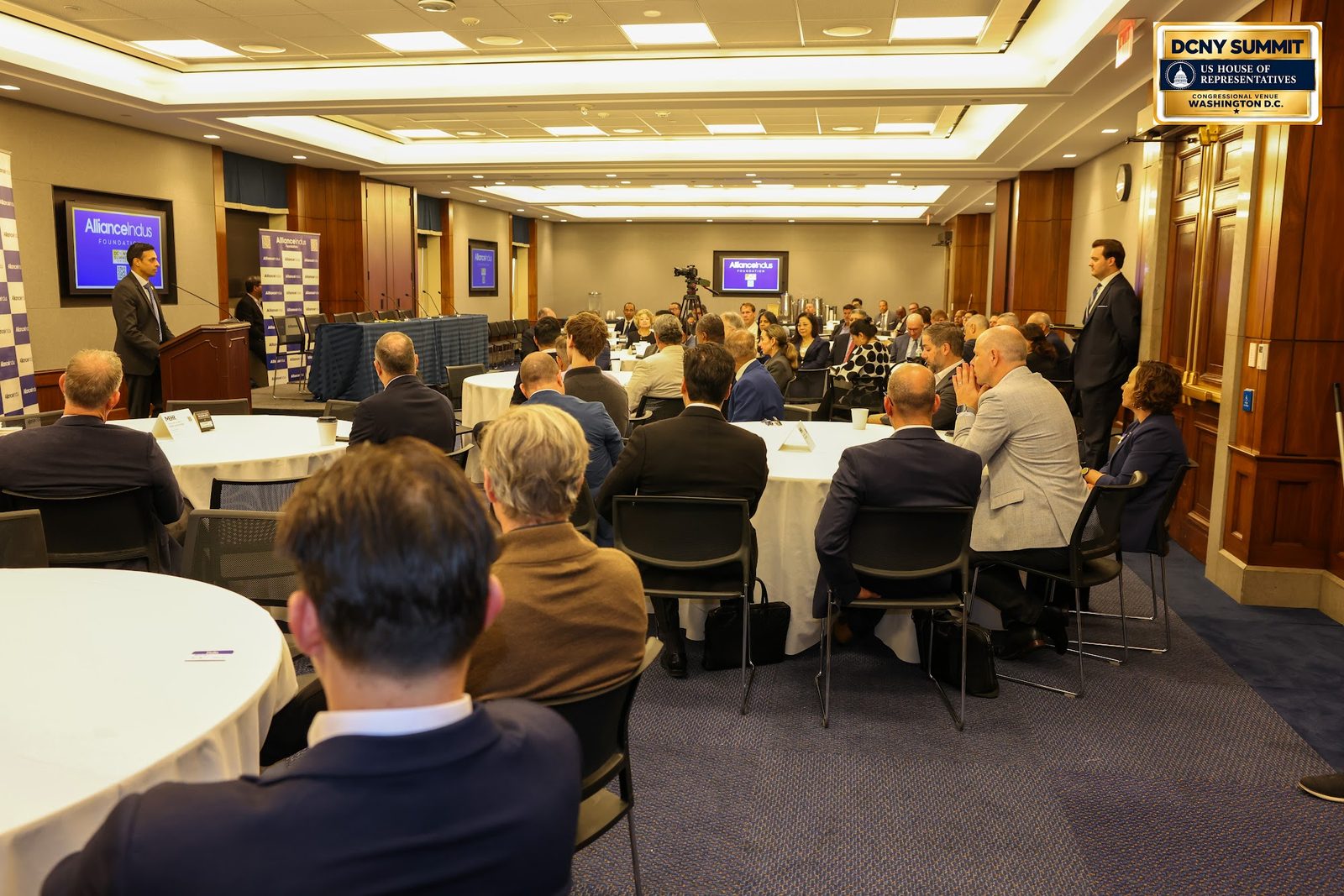 A wide view of the conference room, with guests at round tables and a speaker at the podium in the distance