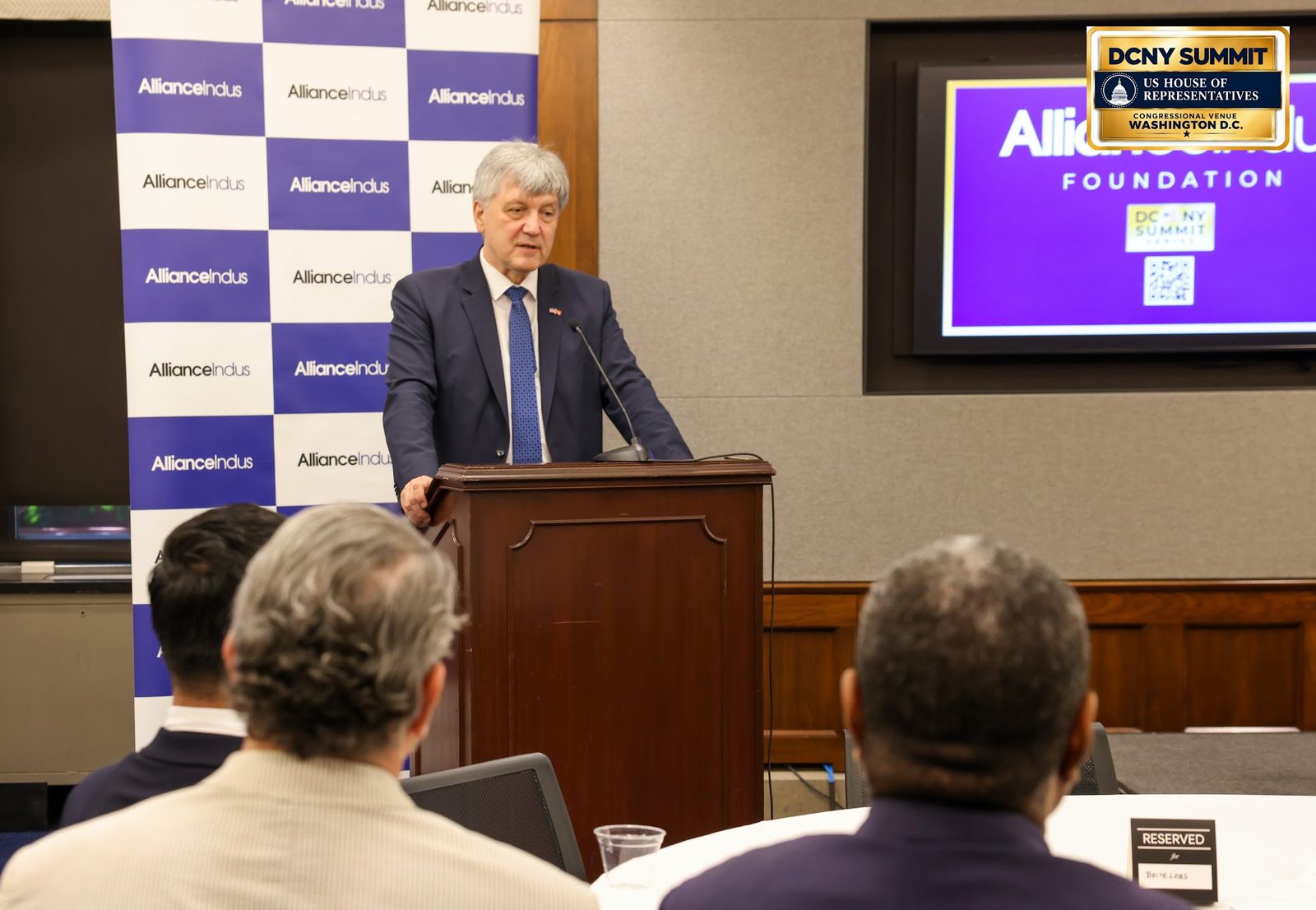Montenegro's ambassador speaks at a wooden podium as guests listen, with an AllianceIndus screen behind