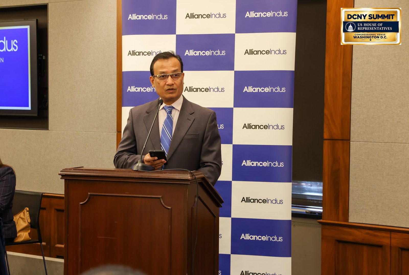 Bangladesh's ambassador reads remarks from a phone at the podium, in front of an AllianceIndus backdrop