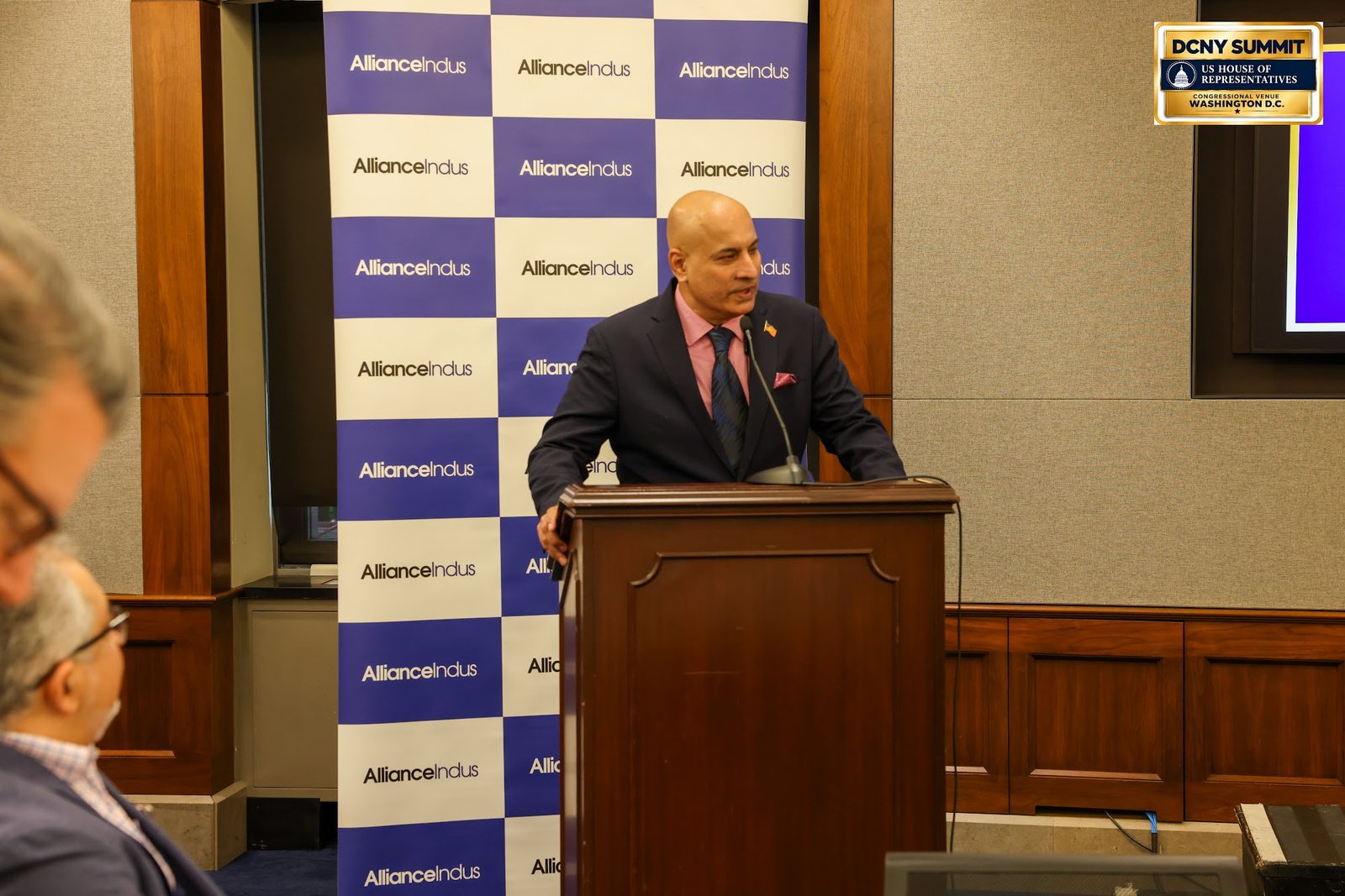 A speaker in a dark suit and red tie at a wooden podium in front of an AllianceIndus backdrop