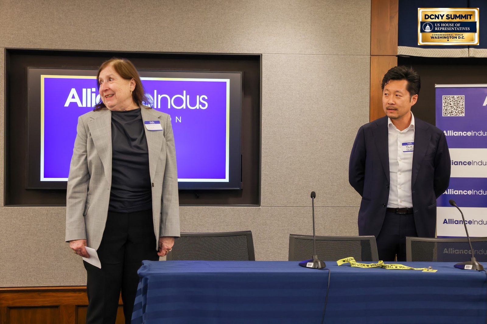 Two speakers standing at the front of the room beside a panel table and an AllianceIndus screen