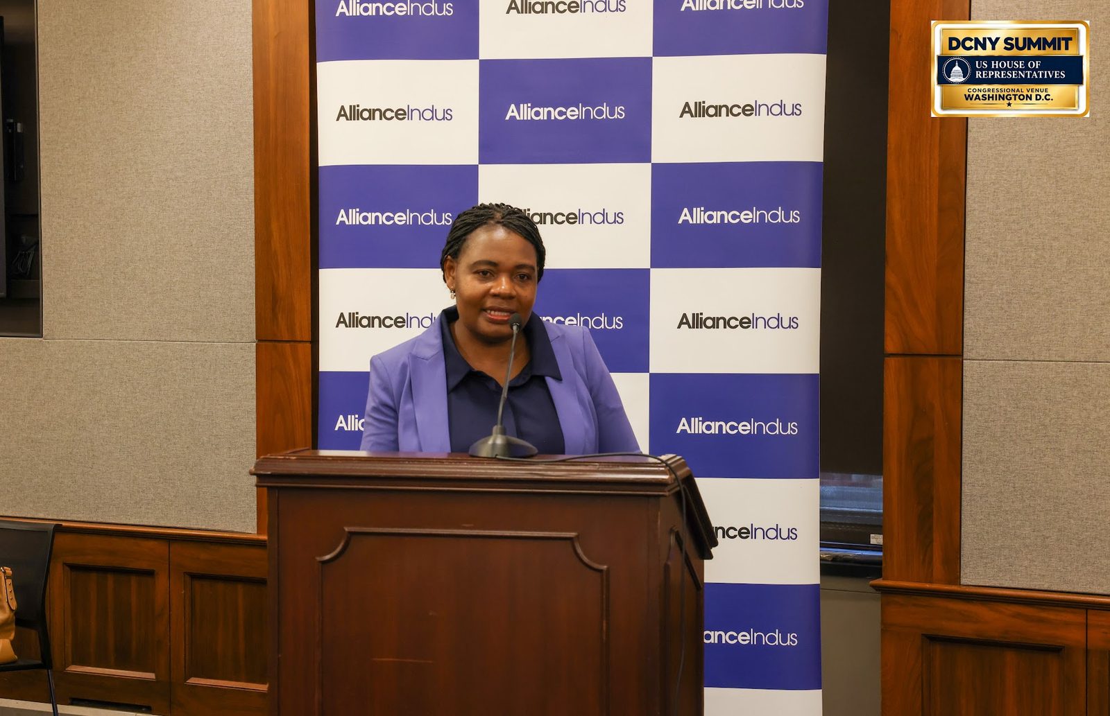 A speaker in a purple jacket at a wooden podium in front of an AllianceIndus backdrop