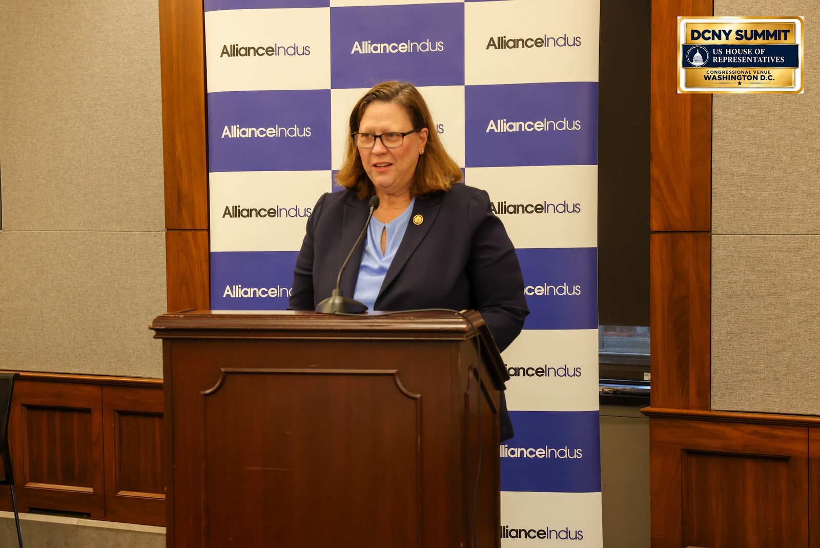 A speaker wearing glasses and a dark blazer at a wooden podium in front of an AllianceIndus backdrop