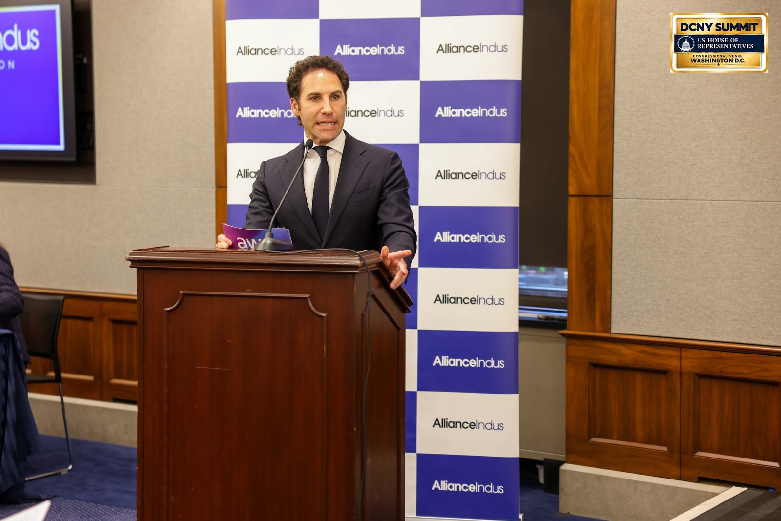 Michael Greenwald speaks at the podium in front of an AllianceIndus backdrop