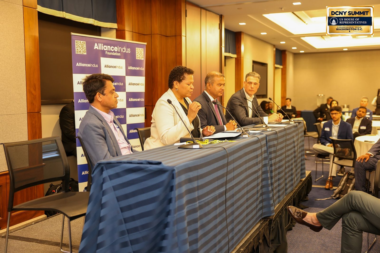 Four panelists at a draped table along the side of the room, with guests seated beyond them