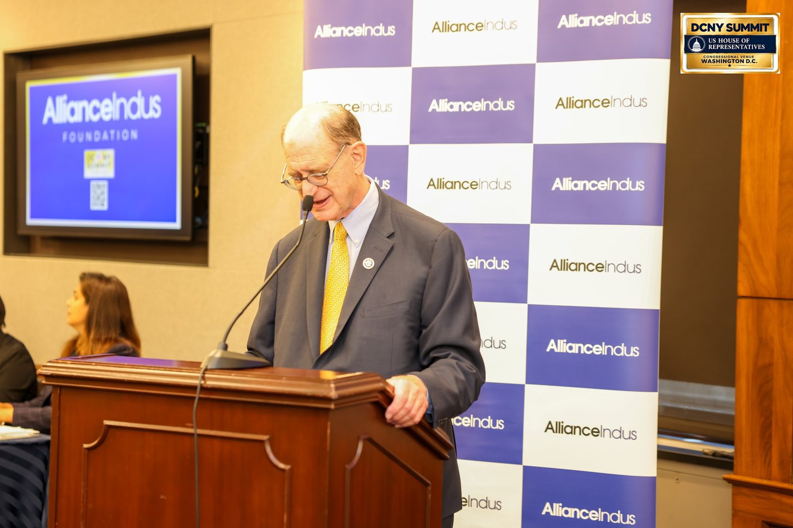A speaker in a dark suit and yellow tie reads remarks at a wooden podium in front of the AllianceIndus backdrop