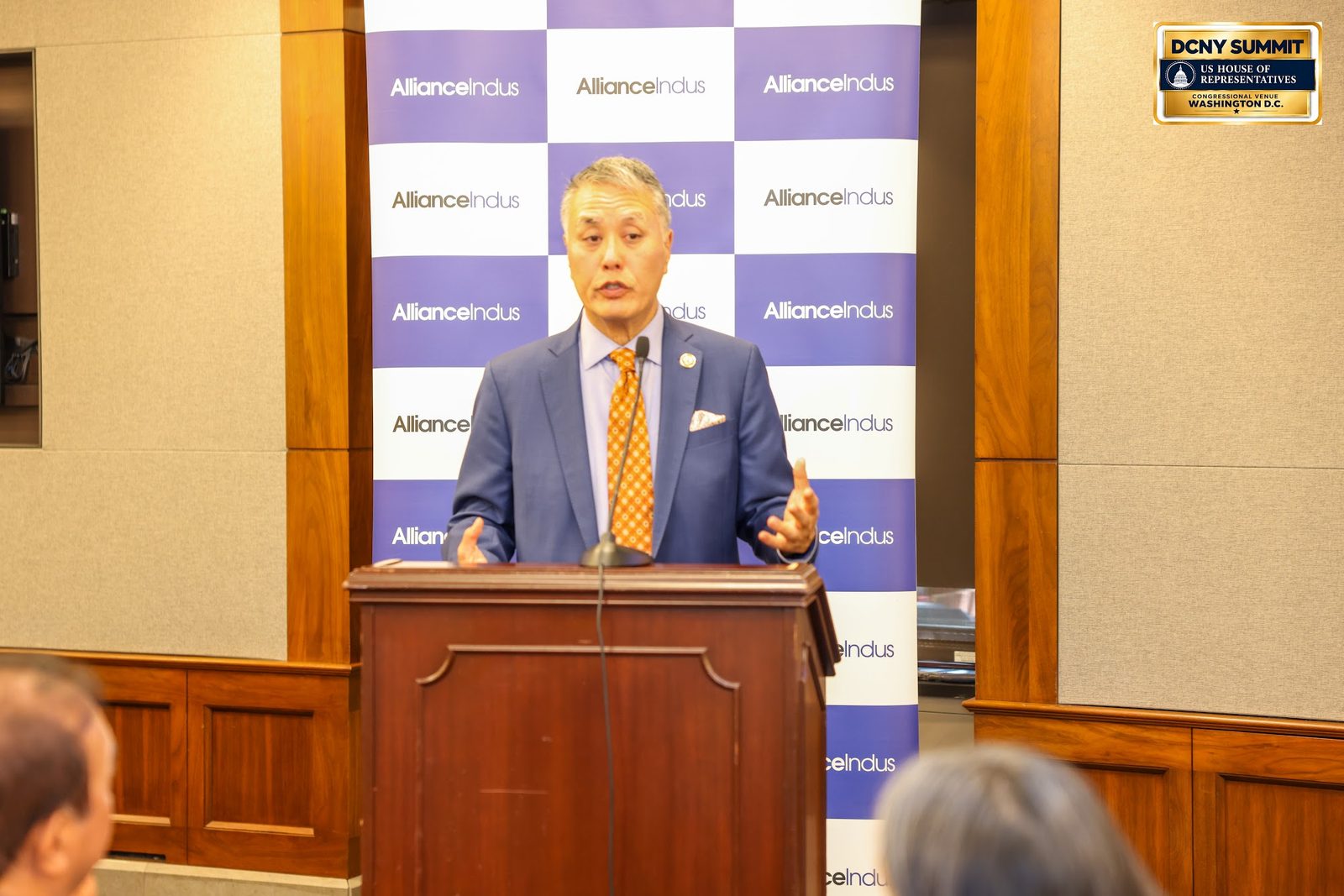 Rep. Mark Takano, in an orange tie, speaks at the podium in front of an AllianceIndus backdrop