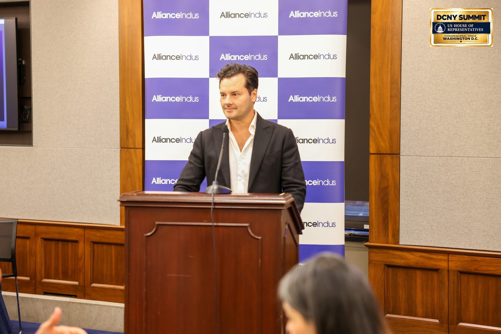 A speaker in a dark jacket at a wooden podium in front of the AllianceIndus backdrop