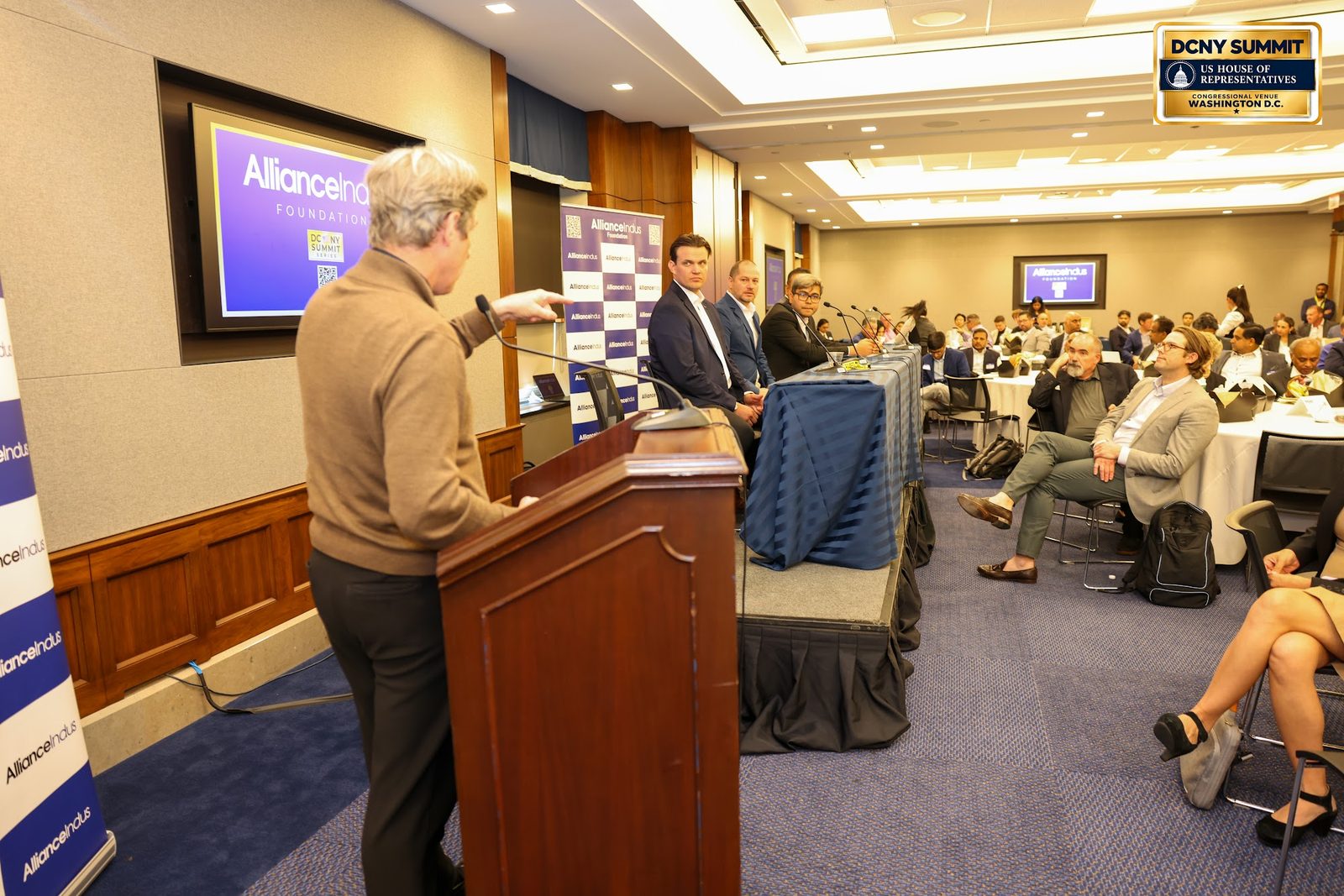 A speaker in a brown sweater at a podium, seen from the side, facing a full room with a panel table behind