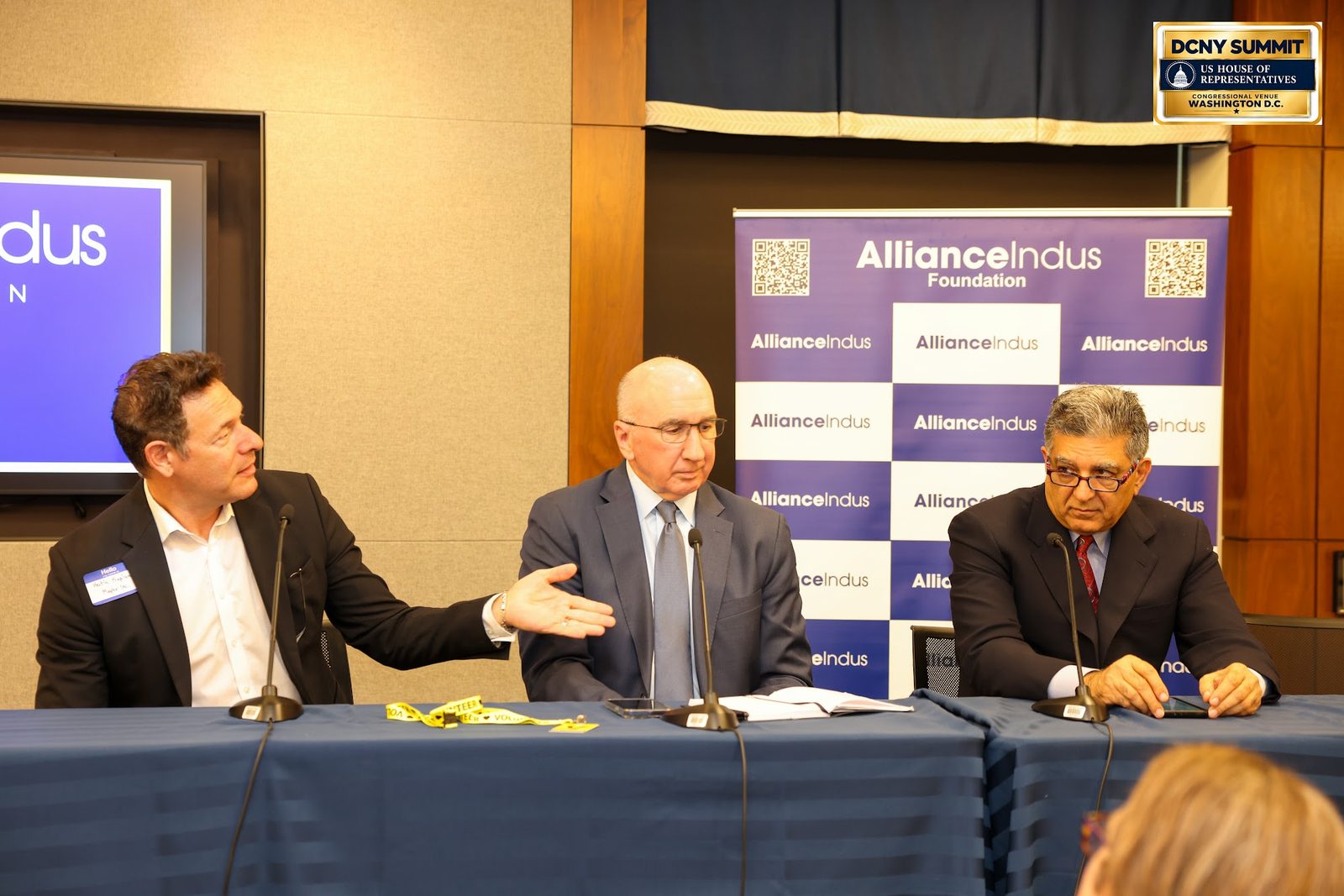 Three panelists at a draped table, one gesturing mid-sentence, in front of an AllianceIndus banner