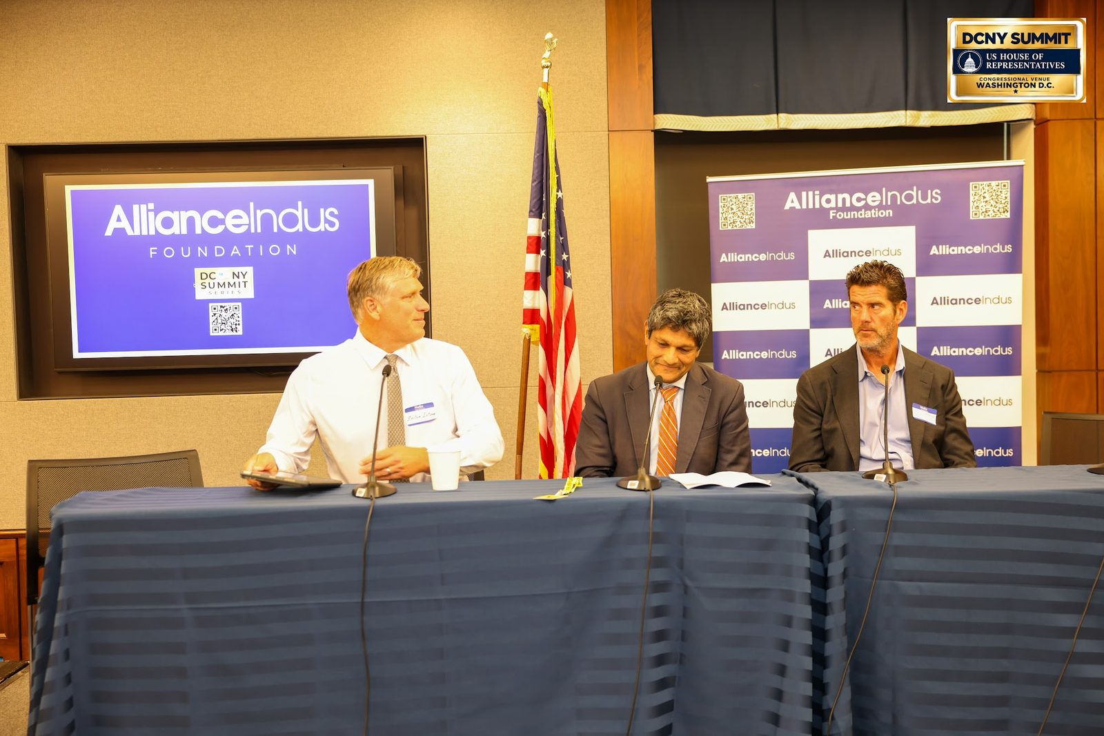 Three panelists at a draped table, with an American flag and AllianceIndus backdrops behind them