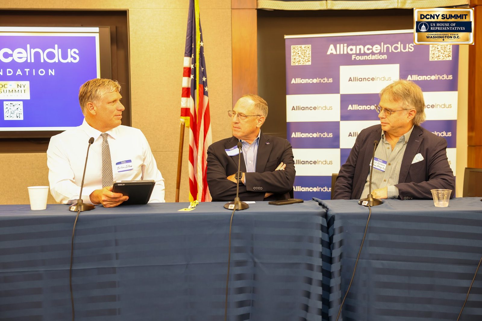 Three panelists at a draped table, one holding a tablet, with an American flag and AllianceIndus backdrops behind them