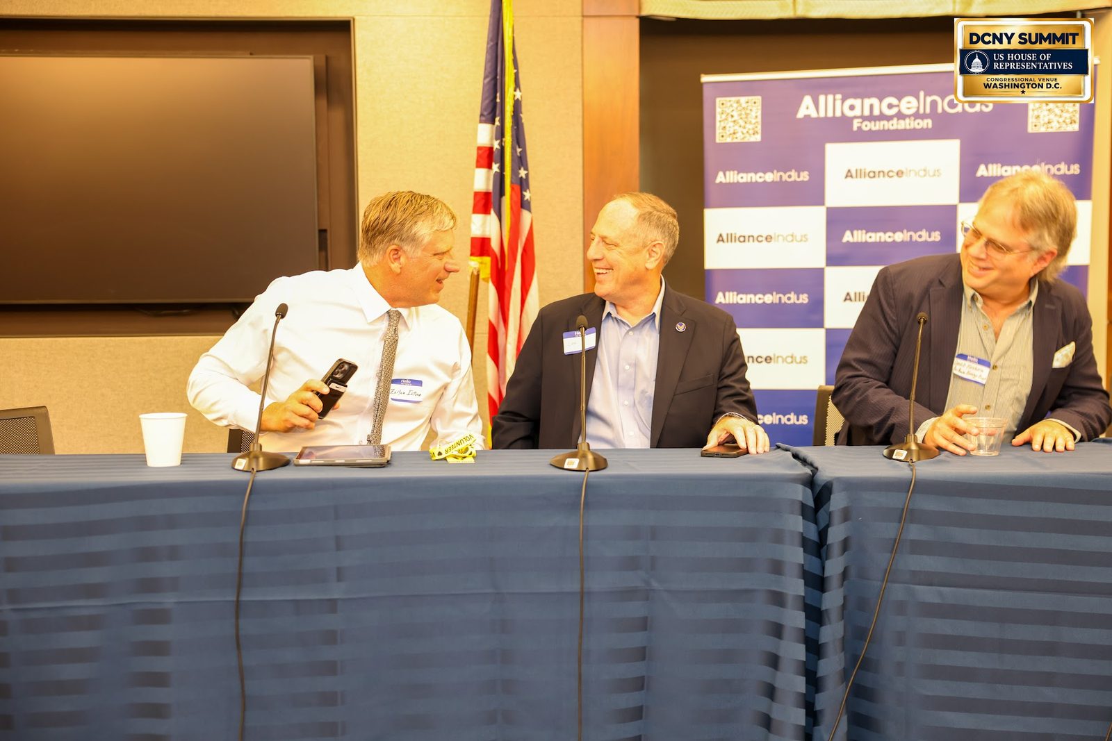 Three panelists seated at a draped table, with the AllianceIndus banner and the U.S. flag behind them