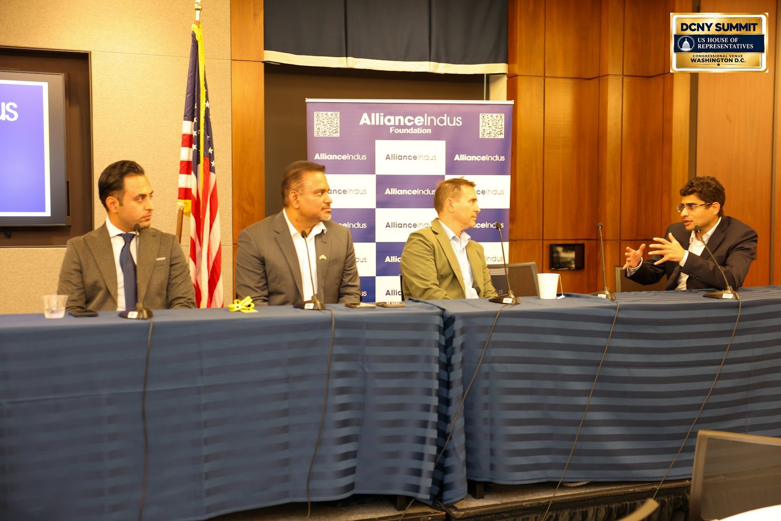 Four panelists seated at a draped table beside the U.S. flag, with the AllianceIndus banner behind them