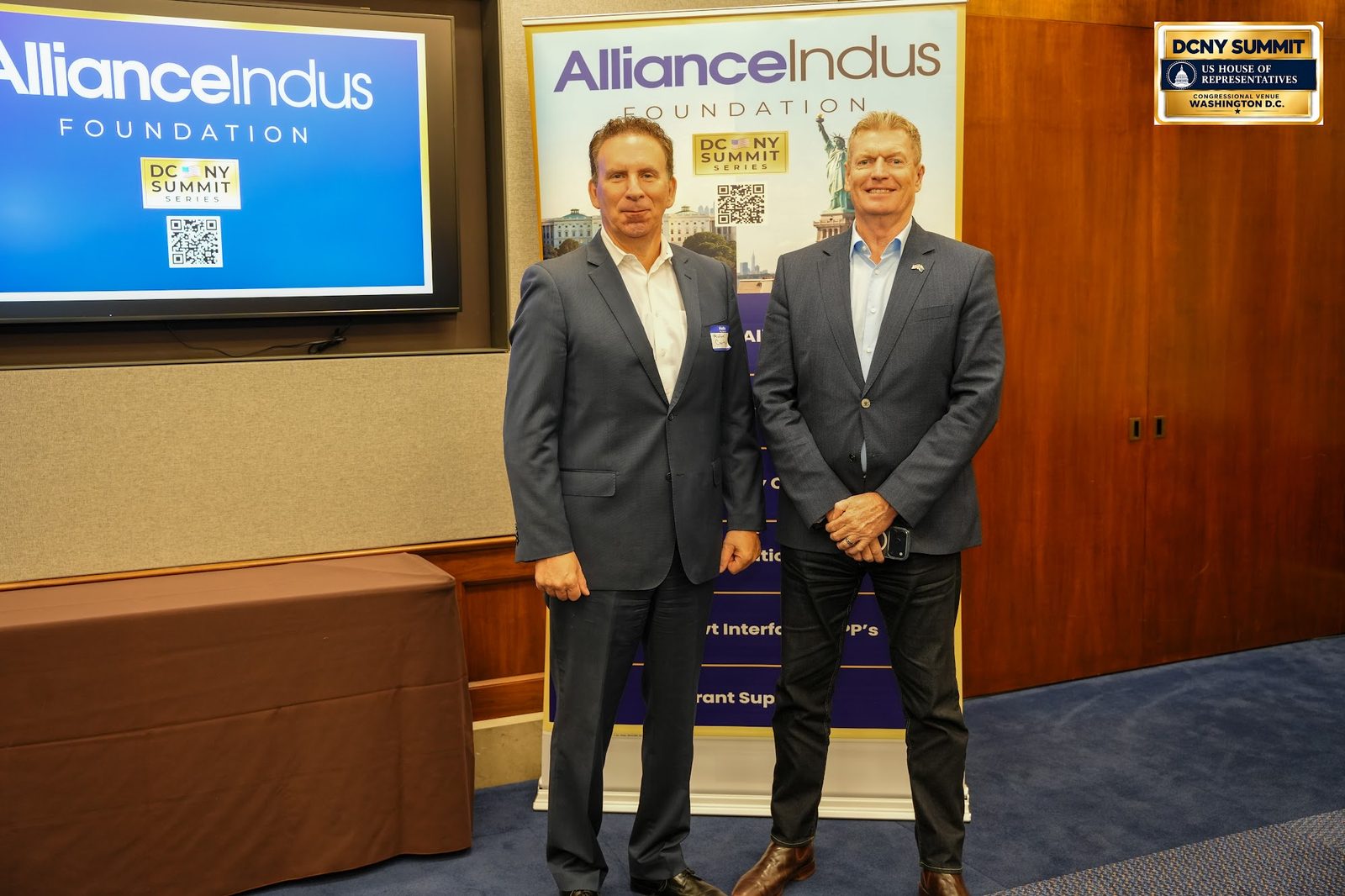 Two men pose in front of a screen showing the AllianceIndus Foundation logo