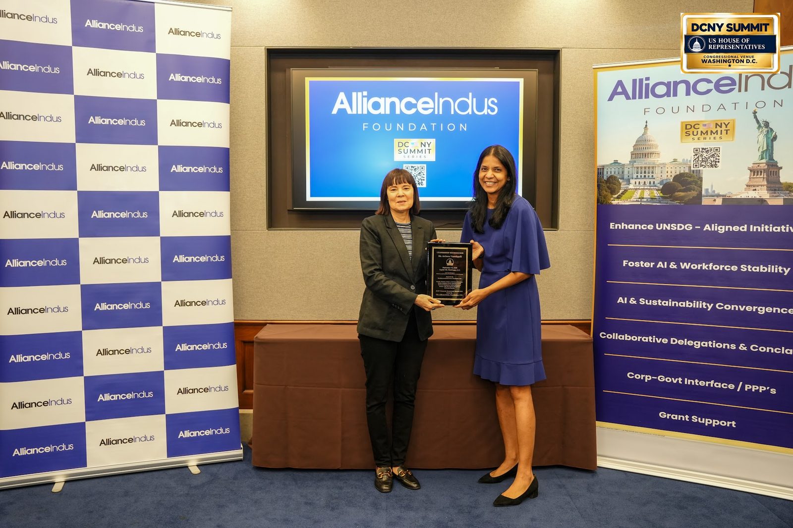 Two women at the AllianceIndus photo wall, one holding a plaque