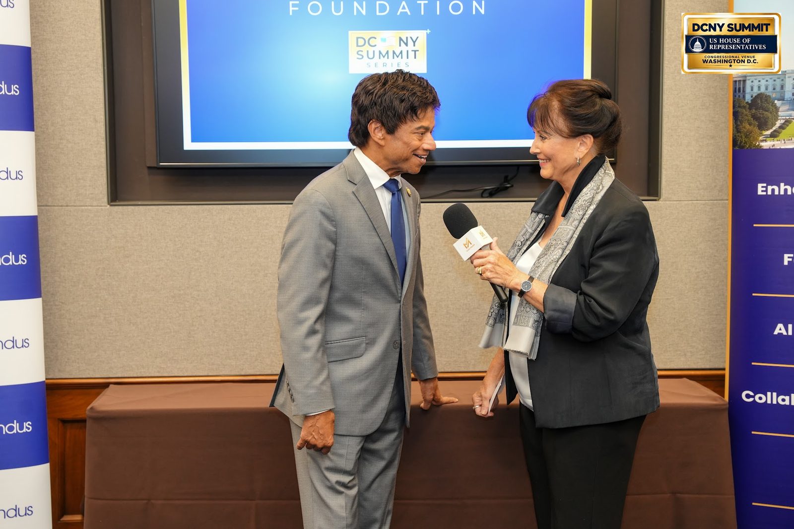 A woman with a microphone interviews a man in a grey suit in front of the AllianceIndus screen