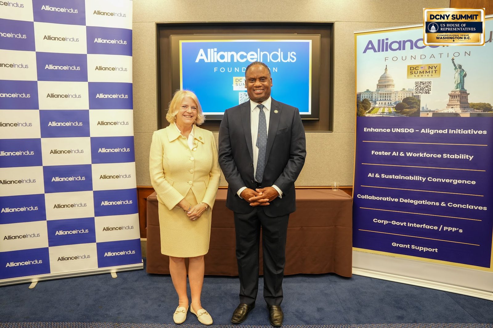 A woman in a pale yellow suit and a man in a dark suit pose at the AllianceIndus photo wall