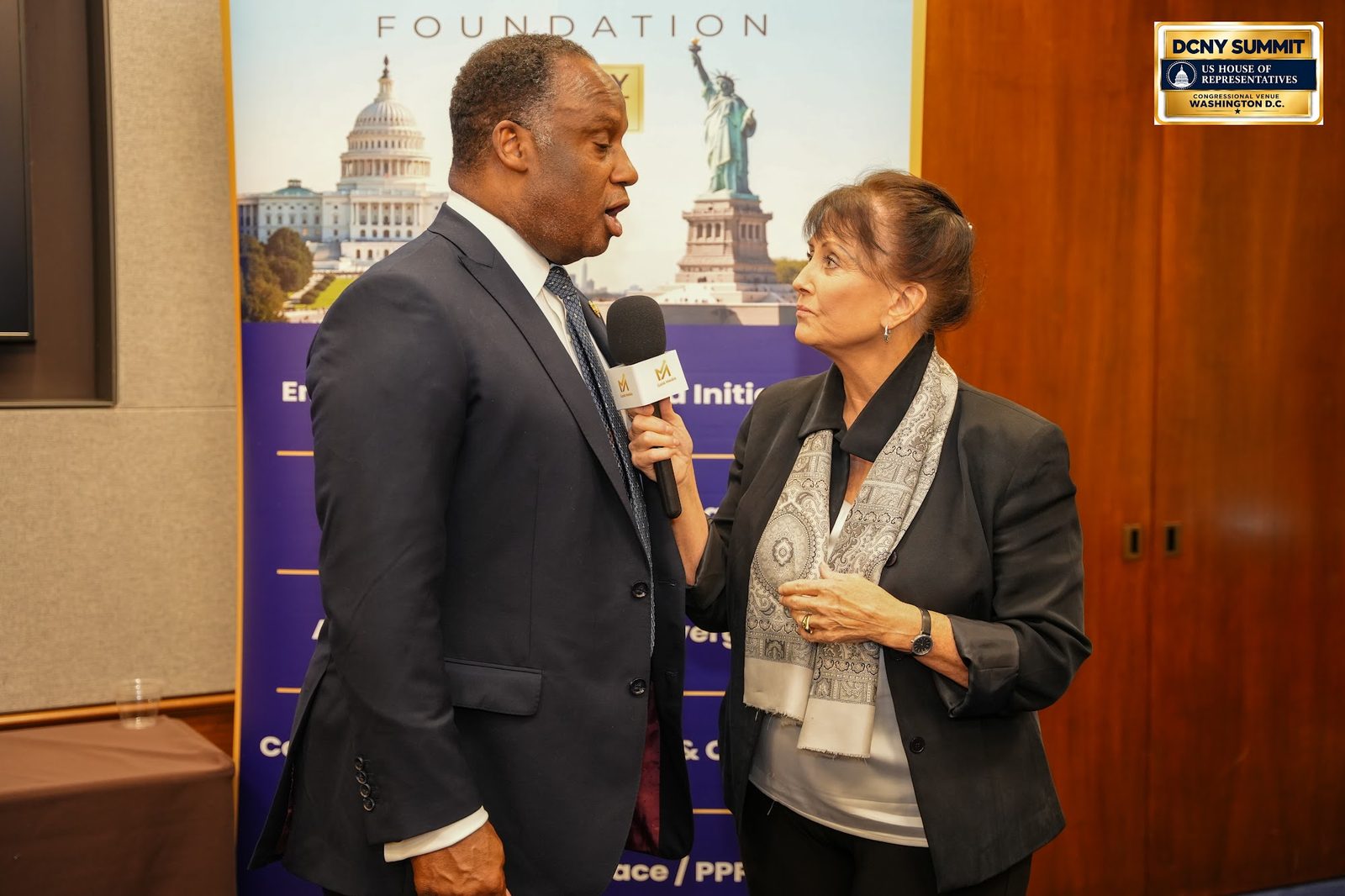 A woman with a microphone interviews a man in a dark suit beside the AllianceIndus banner